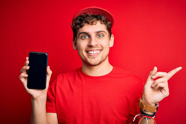 Jovem homem de entrega loira com cabelo encaracolado usando tampa segurando smartphone sobre fundo vermelho muito feliz apontando com a mão e o dedo para o lado
 - Foto, Imagem