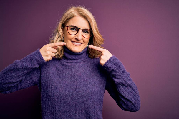 Mujer rubia hermosa de mediana edad vistiendo suéter casual cuello alto púrpura y gafas sonriendo alegre mostrando y señalando con los dedos los dientes y la boca. Concepto de salud dental
. - Foto, Imagen