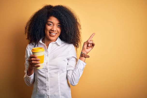 Jonge Afro-Amerikaanse zakenvrouw met afro haar drinken van koffie uit take away cup erg blij wijzend met hand en vinger naar de zijkant - Foto, afbeelding