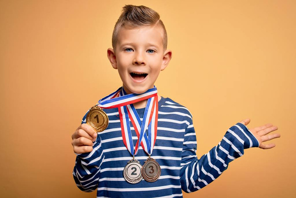 黄色の背景に賞のメダルを身に着けている若い白人の勝者の子供は非常に幸せと興奮,大きな笑顔で叫んで勝利を祝う勝者式と手を上げ - 写真・画像