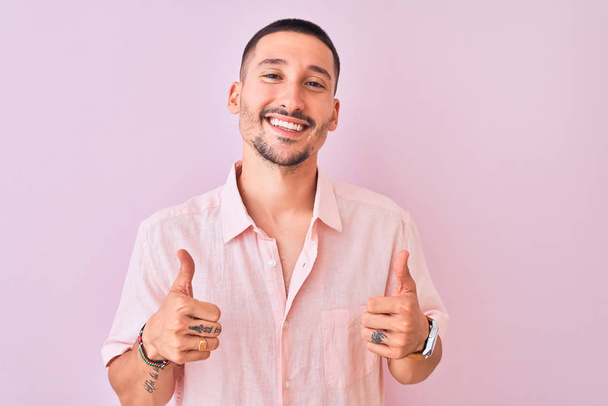 Jonge knappe man draagt roze shirt staan over geïsoleerde achtergrond succes teken doen positieve gebaar met de hand, duimen omhoog glimlachend en gelukkig. Vrolijke uitdrukking en winnaar gebaar. - Foto, afbeelding