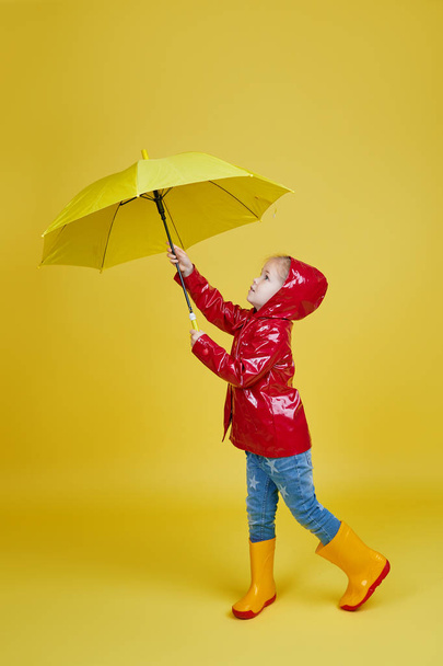 Cheerful child girl with yellow umbrella and red rain coat on colored yellow background. Copy space for text - Фото, изображение