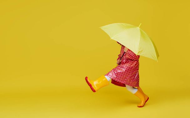 Cheerful child girl with yellow umbrella and red rain coat on colored yellow background. Copy space for text - Foto, afbeelding
