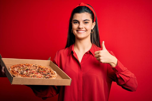 Fiatal gyönyörű barna nő gazdaság szállítási doboz olasz pizza felett piros háttér boldog nagy mosollyal csinál oké jel, hüvelykujj fel ujjaival, kiváló jel - Fotó, kép