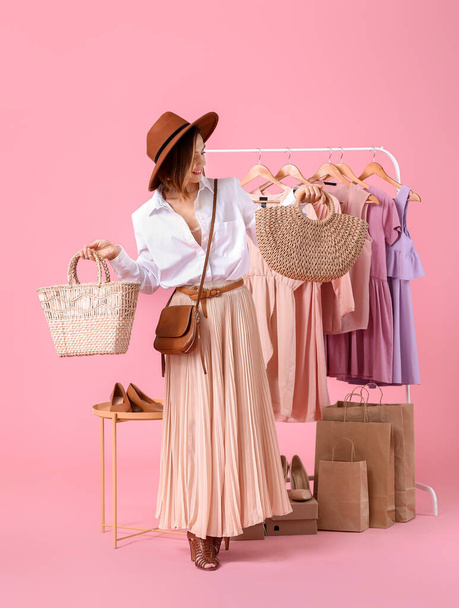 Female stylist with bags and modern clothes on rack against color background - 写真・画像