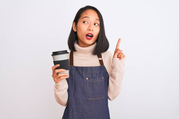 Красивая бариста китайская женщина в фартуке держа кофе на изолированном белом фоне удивлен идеей или вопрос указывая пальцем со счастливым лицом, номер один
 - Фото, изображение