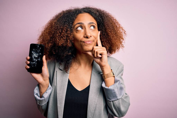 Молодая африканская американка с афроволосами держа треснутый и сломанный экран смартфона серьезное лицо думая о вопросе, очень запутанная идея
 - Фото, изображение