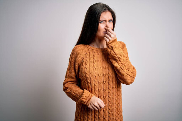 Νεαρή όμορφη γυναίκα με μπλε μάτια που φοράει ένα απλό πουλόβερ που στέκεται πάνω από λευκό φόντο μυρίζοντας κάτι βρωμερό και αηδιαστικό, ανυπόφορη μυρωδιά, κρατώντας την αναπνοή με τα δάχτυλα στη μύτη. Άσχημη μυρωδιά. - Φωτογραφία, εικόνα