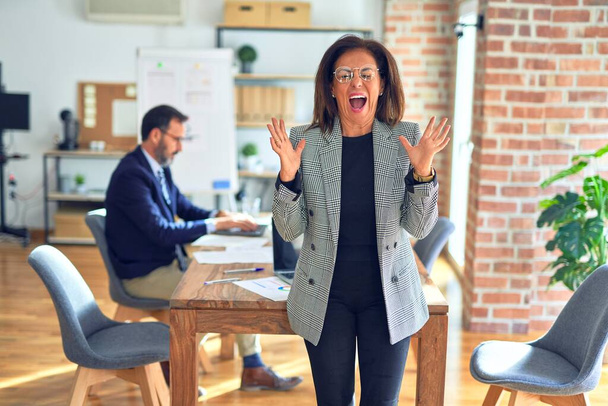 Middle age beautiful businesswoman wearing jacket and glasses standing at the office celebrating mad and crazy for success with arms raised and closed eyes screaming excited. Winner concept - Fotoğraf, Görsel