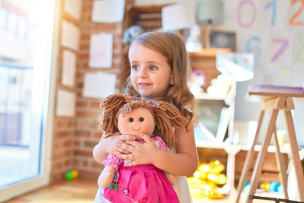 Urocza blond maluch przytulić lalkę wokół wielu zabawek w przedszkolu - Zdjęcie, obraz