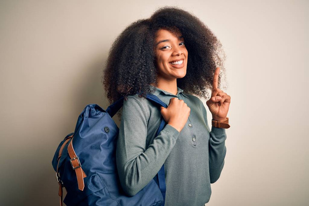 Młoda afrykańska studentka z afro włosami w plecaku na tle zaskoczona pomysłem lub pytaniem wskazującym palcem na szczęśliwą twarz, numer jeden - Zdjęcie, obraz