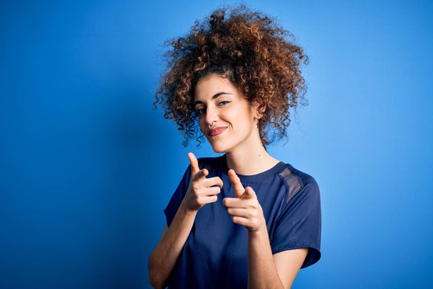 Young beautiful woman with curly hair and piercing wearing casual blue t-shirt pointing fingers to camera with happy and funny face. Good energy and vibes. - Fotó, kép