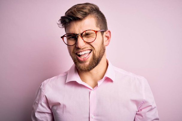 Young handsome blond man with beard and blue eyes wearing pink shirt and glasses winking looking at the camera with sexy expression, cheerful and happy face. - Photo, Image
