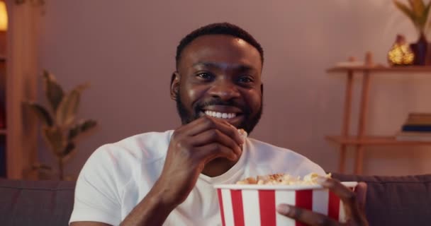 Close up of happy young man sitting on sofa and eating popcorn while enjoing weekend. Bearded guy watching funny show and laughing while watching TV at home. Concept of leisure. - Séquence, vidéo