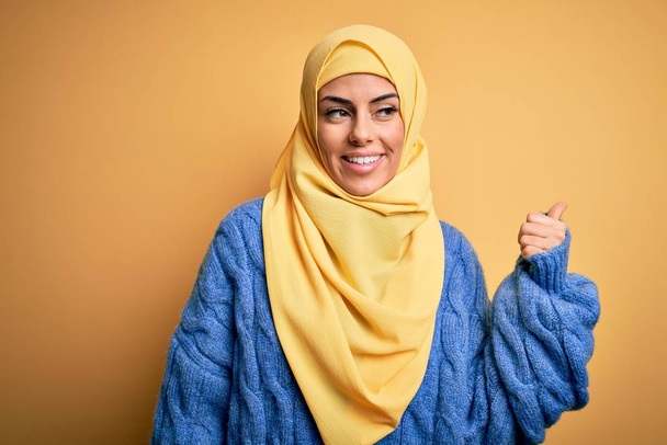 Junge schöne brünette Muslimin trägt arabischen Hijab über isoliertem gelben Hintergrund, lächelt mit glücklichem Gesicht und zeigt mit erhobenem Daumen zur Seite. - Foto, Bild