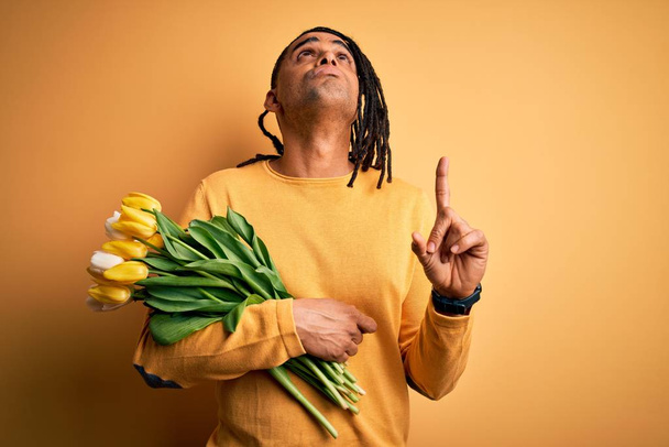 Joven afroamericano afro romántico hombre con rastas sosteniendo ramo de tulipanes amarillos apuntando hacia arriba mirando triste y molesto, indicando la dirección con los dedos, infeliz y deprimido
. - Foto, Imagen