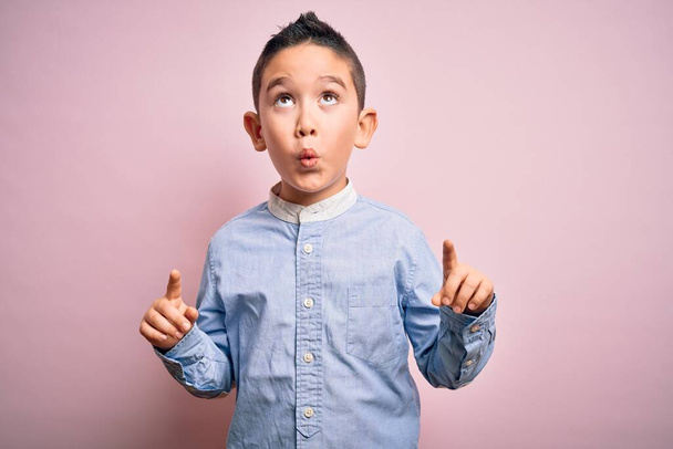 Nuori poika lapsi yllään tyylikäs paita seisoo vaaleanpunainen eristetty tausta hämmästynyt ja yllättynyt etsii ylös ja osoittaa sormilla ja nosti kädet
. - Valokuva, kuva