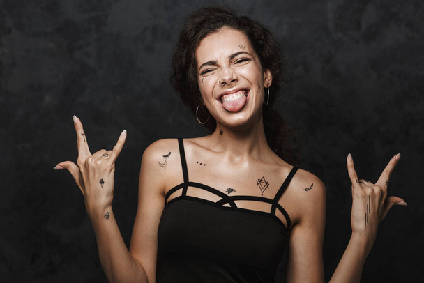 Image of young excited woman with tattoo sticking out her tongue and gesturing horns with fingers isolated over black background - Foto, Imagem