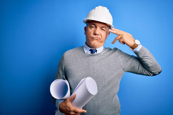 Middle age handsome grey-haired architect man wearing safety helmet holding blueprints Shooting and killing oneself pointing hand and fingers to head like gun, suicide gesture. - Foto, afbeelding