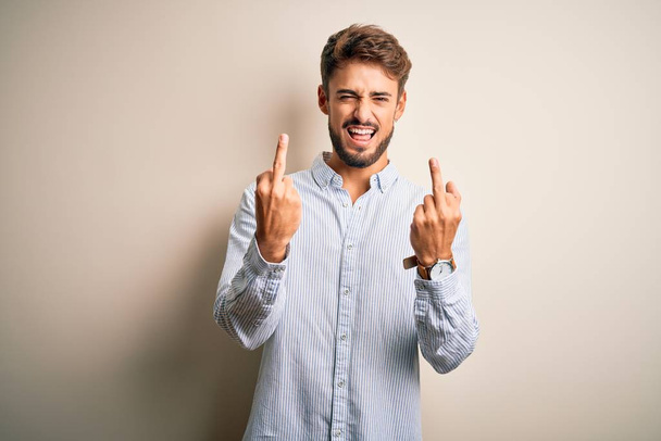 Young handsome man with beard wearing striped shirt standing over white background Showing middle finger doing fuck you bad expression, provocation and rude attitude. Screaming excited - Foto, Bild