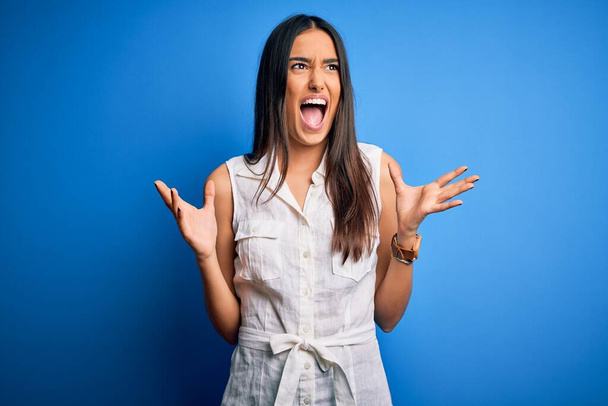 Young beautiful brunette woman wearing casual dress over isolated blue background crazy and mad shouting and yelling with aggressive expression and arms raised. Frustration concept. - Foto, Bild