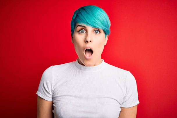 Young beautiful woman with blue fashion hair wearing casual t-shirt over red background afraid and shocked with surprise and amazed expression, fear and excited face. - Photo, Image