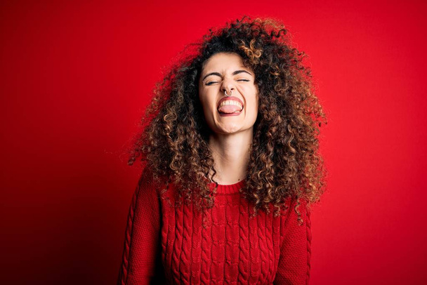 Jonge mooie vrouw met krullend haar en piercing dragen casual rode trui steken tong uit blij met grappige uitdrukking. Emotieconcept. - Foto, afbeelding