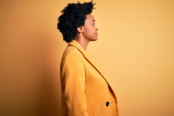 Νεαρή όμορφη Αφρο-Αμερικανίδα επιχειρηματίας με σγουρά μαλλιά φορώντας κίτρινο μπουφάν κοιτάζοντας προς τα πλάγια, χαλαρώστε προφίλ ποζάρουν με φυσικό πρόσωπο με αυτοπεποίθηση χαμόγελο. - Φωτογραφία, εικόνα
