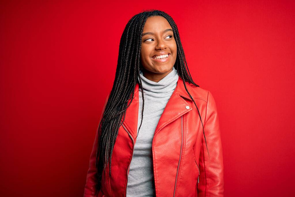 Junge Afroamerikanerin in cooler modischer Lederjacke über rotem isoliertem Hintergrund, die mit einem Lächeln im Gesicht und natürlichem Gesichtsausdruck zur Seite schaut. Zuversichtliches Lachen. - Foto, Bild