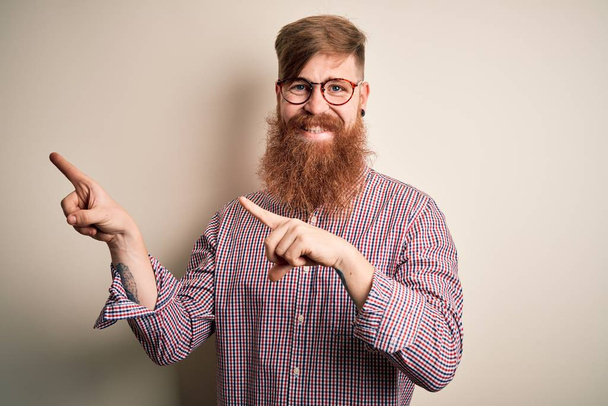 ハンサムなアイルランドの赤毛ビジネス男あります髭を身に着けています眼鏡上の隔離された背景笑顔と見てカメラを指しています二つの手と指で側面. - 写真・画像