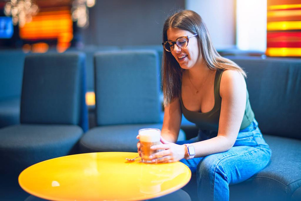 Young beautiful woman smiling happy and confident. Sitting with smile on face holding glass of coffee at restaurant - Foto, Imagen