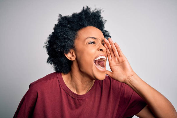 若い美しいアフリカ系アメリカ人のアフロ女性は、カジュアルなTシャツを着て大声で叫び、口の上に手で横に大声で叫んで立って巻き毛。コミュニケーションの概念. - 写真・画像