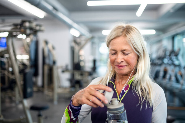 A senior woman with earphones and water bottle standing in gym. - Foto, Imagen