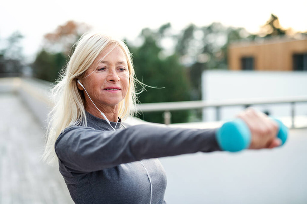 A senior woman with earphones and dumbbells outdoors doing exercise. - Foto, immagini