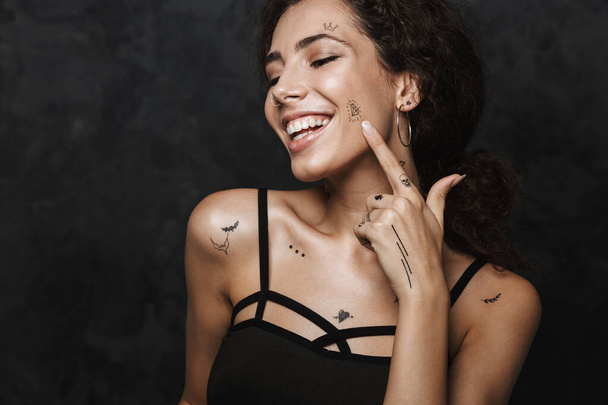 Immagine di una donna riccia felice che sorride e punta il dito al suo tatuaggio isolato su sfondo nero
 - Foto, immagini