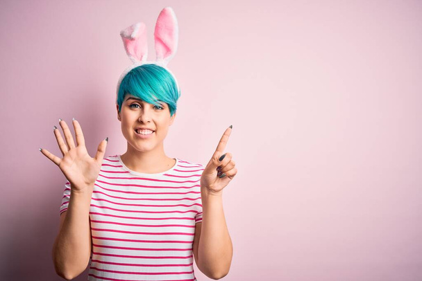 Młoda kobieta z modą niebieskie włosy noszące wielkanocne uszy królika nad różowym tle pokazując i wskazując palcem numer sześć uśmiechając się pewny siebie i szczęśliwy. - Zdjęcie, obraz