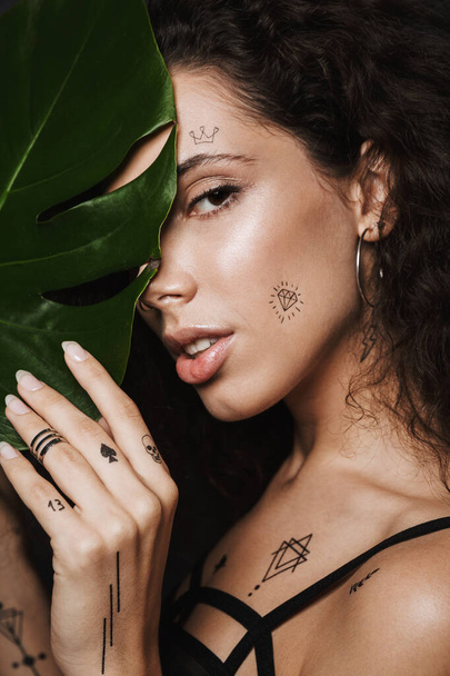 Image of young seductive woman with tattoo looking at camera behind green leaf isolated over black background - Photo, image