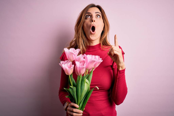 Junge schöne brünette Frau mit einem Strauß rosa Tulpen vor isoliertem Hintergrund erstaunt und überrascht nach oben schauend und mit erhobenen Fingern und Armen zeigend. - Foto, Bild