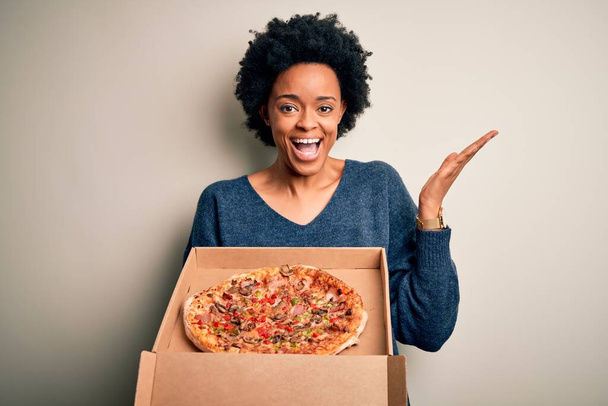 Jonge Afro-Amerikaanse afro vrouw met krullend haar holding delivery box met Italiaanse pizza erg blij en opgewonden, winnaar uitdrukking vieren overwinning schreeuwen met een grote glimlach en opgeheven handen - Foto, afbeelding