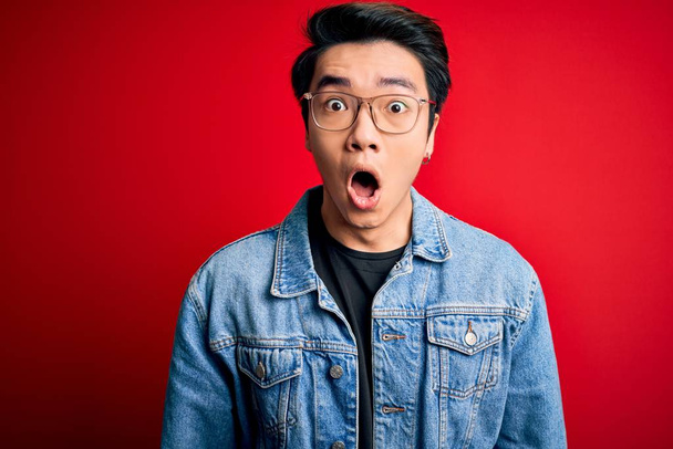 Молодой красивый китайский мужчина в джинсовой куртке и очках на красном фоне боится и шокирован удивлением выражения лица, страха и возбужденного лица
. - Фото, изображение