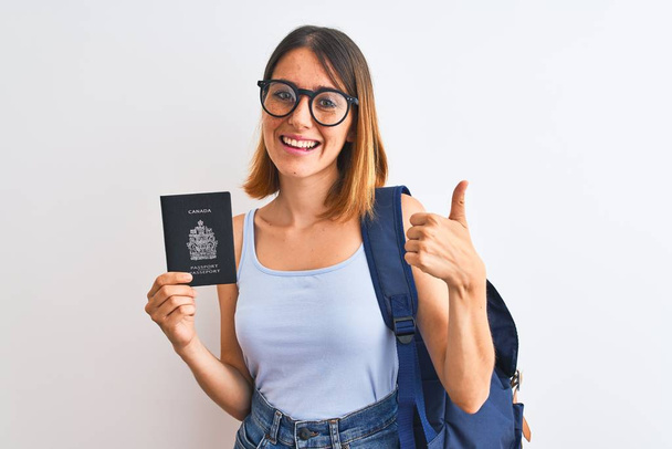 Mulher estudante ruiva bonita vestindo mochila e segurando passaporte do canadá feliz com grande sorriso fazendo sinal ok, polegar com os dedos, excelente sinal
 - Foto, Imagem