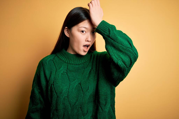 Молода красива азіатка в зеленому зимовому светрі над жовтим ізольованим тлом, здивована рукою на голові за помилку, пам'ятає помилку. Забули, погана концепція пам'яті
. - Фото, зображення