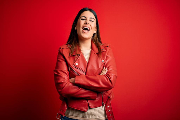 Young beautiful woman wearing red leather jacket over isolated background happy face smiling with crossed arms looking at the camera. Positive person. - Photo, Image