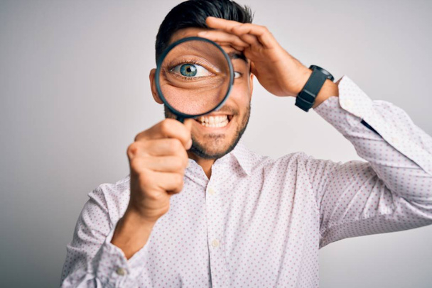 Young detective man looking through magnifying glass over isolated background stressed with hand on head, shocked with shame and surprise face, angry and frustrated. Fear and upset for mistake. - Photo, Image