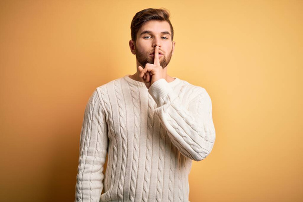 Young blond man with beard and blue eyes wearing white sweater over yellow background asking to be quiet with finger on lips. Silence and secret concept. - Foto, immagini