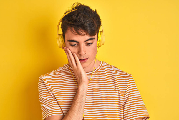 Adolescente niño usando auriculares sobre fondo amarillo aislado pensando que se ve cansado y aburrido con problemas de depresión con los brazos cruzados
. - Foto, Imagen