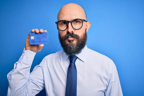 Przystojny łysy biznesmen z brodą trzymający kartę kredytową na odizolowanym niebieskim tle z pewnym siebie wyrazem mądrej twarzy myśląc poważnie - Zdjęcie, obraz