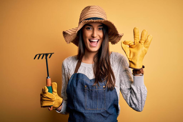 若い美しい農家の女性を身に着けているエプロンと帽子を使用してください指でokサインを行う黄色の背景に熊手,優れたシンボル - 写真・画像