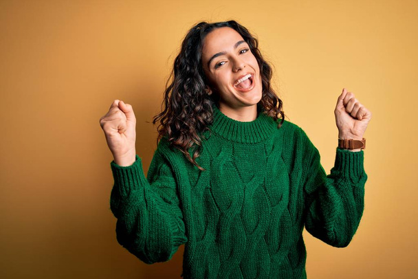 Young beautiful woman with curly hair wearing green casual sweater over yellow background celebrating surprised and amazed for success with arms raised and open eyes. Winner concept. - Photo, Image