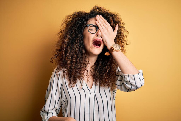 Young beautiful woman with curly hair and piercing wearing striped shirt and glasses Yawning tired covering half face, eye and mouth with hand. Face hurts in pain. - Photo, Image
